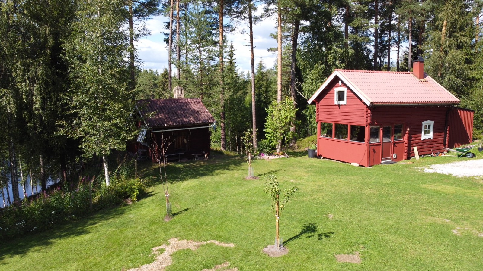 The red timber house and an older outbuilding on a mown lawn, forest and the river just behind