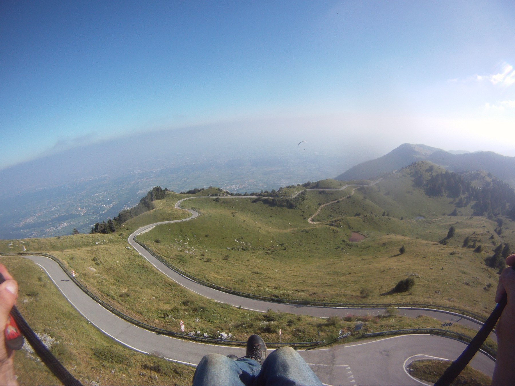 Point-of-view from a paraglider harness, feet dangling above a hairpin mountain road